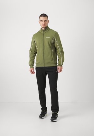Chaqueta de forro polar verde con cremalleras y ribetes blancos, combinada con pantalones negros y zapatillas negras con acentos verdes.