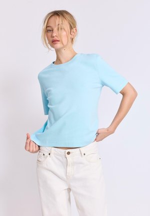 Žena s blond vlasmi, oblečená v svetlomodrej tričku s krátkym rukávom a smotanových nohaviciach, drží lem trička a pózuje pred jednofarebným pozadím.