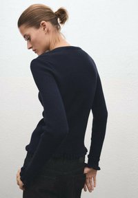 Suéter de canalé azul marino con mangas largas, dobladillo con volantes y tela texturizada, combinado con jeans negros sobre un fondo claro.