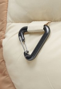 Czarny trójkątny karabinek przypięty do beżowego paska z tkaniny na kremowej, wyściełanej powierzchni.