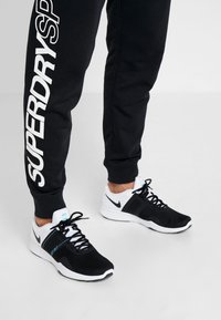 Schwarze Sportschuhe mit weißen Akzenten und einem strukturierten Mesh-Obermaterial, kombiniert mit schwarzen Jogginghosen mit weißem Logostin.