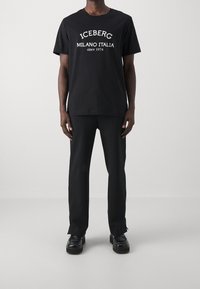 Musta puuvillapaita, jossa on valkoiset tekstit "ICEBERG MILANO ITALIA vuodesta 1974." Yhdistetty mustiin housuihin ja mustiin kenkiin. Yksinkertainen muotoilu.