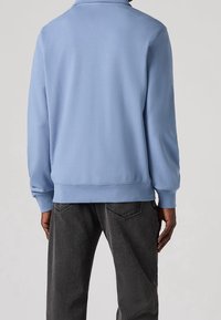 Blå sweatshirt i en bomullsblandning med avslappnad passform, ribbade muddar och fåll. Buren över mörkgrå jeans, vilket visar en jämn textur.