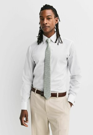 Mann mit Dreadlocks, der ein weißes Hemd, eine hellgraue Krawatte, beige Hose und einen braunen Gürtel trägt, steht vor einem schlichten hellen Hintergrund.
