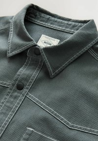 Dark grey collared shirt made of textured material featuring contrasting white stitching, two black buttons, and a chest pocket.