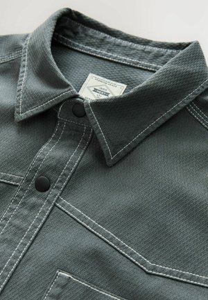 Tumehall collane särk, mis on valmistatud tekstureeritud materjalist ja millel on kontrastne valge õmblus, kaks musta nuppu ja rinna tasku.