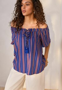 Top a spalle scoperte, a righe nei toni del blu, viola e rosa. Presenta un fiocco con perline e nappine al centro davanti. Tessuto leggero.