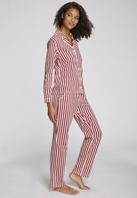 Vrouw die blootvoets staat in een rode en witte verticaal gestreepte lange mouwen pyjama set met een knoopsluiting bovenaan en een broek.