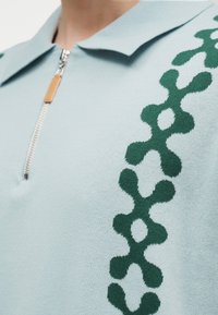 Ανοιχτό μπλε πλεκτό πόλο πουκάμισο με σκούρο πράσινο αφηρημένο σχέδιο αλυσίδας στον ώμο και ασημί φερμουάρ με δερμάτινη ταινία έλξης.