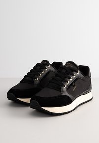 Black sneakers made of mesh and suede, featuring gold eyelets, a cream midsole, textured accents, and a branded logo on the side.