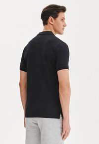 Czarna koszulka polo z krótkim rękawem, klasycznym kołnierzykiem, miękką teksturą i bocznymi rozcięciami. W zestawieniu z szarymi szortami, prezentuje neutralny, casualowy wygląd.