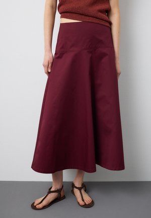 Person trägt einen hoch taillierten, burgunderfarbenen A-Linien-Rock mit einem kurzen, rostfarbenen Pullover und braunen, flachen Sandalen mit Riemchen.