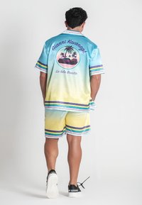 Kurzärmliges, gradient-blaues und gelbes Seidenhemd mit tropischem Druck, kombiniert mit passenden Shorts; lila und grüne Streifen setzen Akzente.