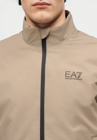 Άνδρας που φοράει μπεζ μπουφάν με φερμουάρ και όρθιο γιακά, με μαύρο λογότυπο EA7 Emporio Armani στο στήθος.