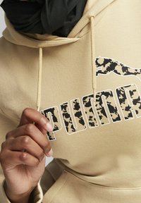 Sudadera beige con un logo 'PUMA' en estampado negro y de camuflaje. Cuenta con capucha ajustable y está hecha de un material suave y texturizado.
