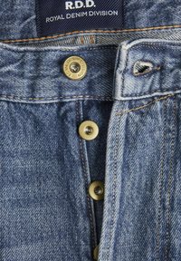 Džínové šortky s modrým prístupom, s mosadznými gombíkmi, zipsom a štítkom s nápisom "R.D.D. Royal Denim Division." Viditeľná textúrovaná tkanina.
