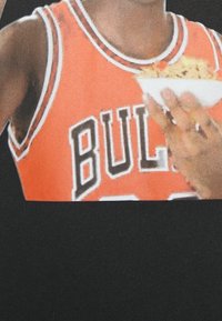 T-shirt noir avec un graphique d'une personne en maillot orange portant le texte "BULLS", tenant un bol de nourriture. Texture lisse, design décontracté.