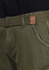 Pantalons vert olive avec une ceinture tressée dotée d'un accent en cuir rectangulaire. Couture visible et tissu texturé.