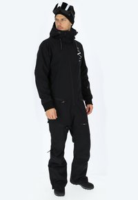 Tuta da neve nera a pezzo unico con chiusura a zip, cappuccio regolabile e molteplici tasche con zip. Realizzata in materiale resistente e impermeabile.