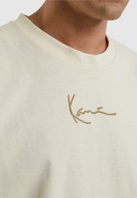 T-shirt en coton de couleur crème avec un col rond. Présente un détail signé brodé marron sur le devant, texture lisse.
