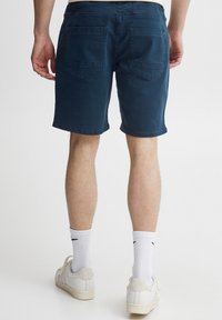 Pantalones cortos de algodón azul marino con corte recto, bolsillos traseros con costuras triangulares y bajos con puños. Llevados con calcetines y zapatos blancos.