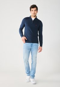 Námořnický modrý svetr na zip s dlouhými rukávy, vyrobený z měkké látky, spárovaný se světle modrými džíny a bílými teniskami; má červené logo.