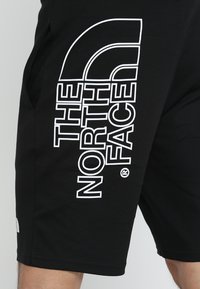 Mustad pehmekoestest mustad lühikesed püksid, mille külgedel on suur valge logo. Disaini kuuluvad taskud ja sirge lõige allservas.