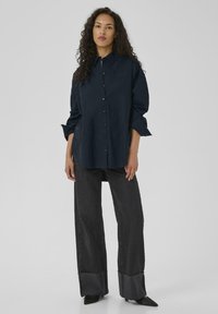 Marineblå knap-op skjorte med krave, parret med brede mørke jeans med flosset kant og spidse sorte sko.