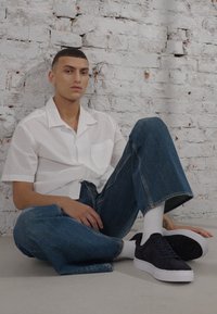 Cămașă albă texturată cu mânecă scurtă, blugi largi de culoare albastră și adidași din suede albastru închis cu tălpi albe, așezat în fața unui fundal din cărămidă albă.