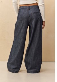Pantalones de mezclilla de pierna ancha en azul oscuro. Presentan una textura suave, detalles cosidos y dos bolsillos en la parte trasera. Un sutil desgastado añade profundidad.