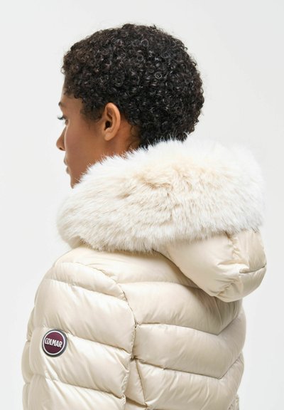 Personne aux cheveux courts, bouclés et noirs portant une veste matelassée couleur crème avec une capuche bordée de fourrure et un écusson du logo Colmar sur la manche.