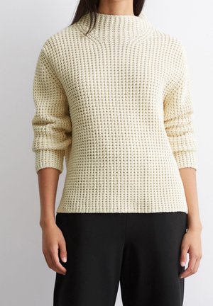 Frau trägt einen cremefarbenen, strukturierten Strickpullover mit hochgekrempelten Ärmeln und schwarze Hose, steht vor einem schlichten Hintergrund.