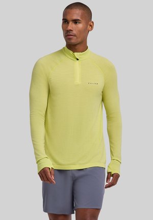 Man wearing a light green long-sleeve zip-up shirt with thumb holes and gray shorts, standing against a plain background.