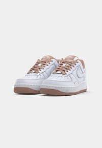 Une paire de baskets Nike Air Force 1 blanches avec des lacets, des semelles et un revêtement intérieur marron, présentée sur un fond blanc uni.