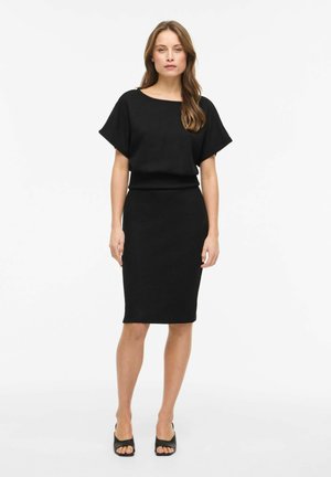 Žena stojaca, oblečená v čiernom tričku s krátkym rukávom a zodpovedajúcej ceruzkovej sukni po kolená, s čiernymi otvorenými lodičkami na bielom pozadí.