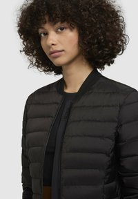 Čierna prešívaná bomber bunda s rebrovaným golierom, predným zipsom a bezšvovým šitím. Ľahký materiál s elegantnou, hladkou textúrou.