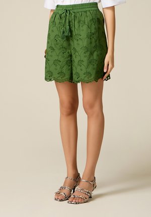 Vrouw draagt groene geborduurde shorts met geschulpte randen en een trekkoord met kwast, gecombineerd met sandalen met open tenen en bandjes met patroon.