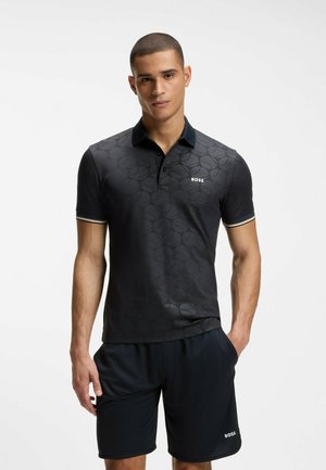 Mand iført sort mønstret polo og sorte shorts, begge med "BOSS" logo, stående med den ene hånd i lommen mod hvid baggrund.