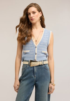 Donna con capelli lunghi e ondulati che indossa un gilet senza maniche lavorato a maglia blu e bianco, jeans a vita alta, cintura larga beige e gioielli dorati.