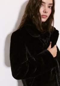Czarny płaszcz z sztucznego futra z szerokim kołnierzem i miękką fakturą, z długimi rękawami i luźnym krojem, częściowo zapięty z przodu.