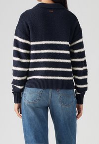 Tmavě modrý pletený svetr s tlustými bílými pruhy, žebrovanými manžetami a lemem. Má kulatý výstřih a malou etiketu na zadní straně.
