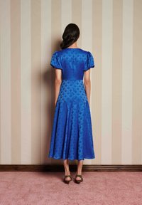 Γυναίκα που στέκεται με την πλάτη στραμμένη προς την κάμερα, φορώντας ένα μπλε μίντι φόρεμα με μοτίβο και φουσκωτά μανίκια, καθώς και τακούνια με λεοπάρ σχέδιο, μπροστά σε τοίχο με ρίγες.