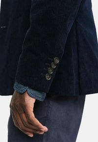 Marineblauer Cord-Blazer mit strukturierter Oberfläche, der vier Knopfdetails an den Bündchen aufweist. Darunter ist der Kragen eines Denim-Hemdes sichtbar.