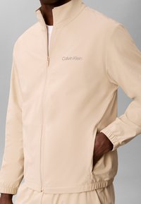 Homme portant une veste beige zippée avec le logo Calvin Klein sur la poitrine, main dans la poche, col de chemise blanche visible en dessous.