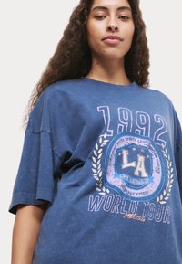 Blue oversized t-shirt with short sleeves featuring a graphic print of "1992" and "LA WORLD TOUR" with laurel design and distressed texture.