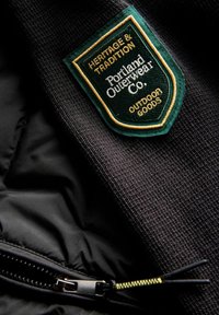 Crna tkanina s teksturiranim završetkom, s zelenim i zlatnim izvezenim oblogom koji kaže "Nasljeđe i tradicija, Portland Outerwear Co." blizu zatvarača.