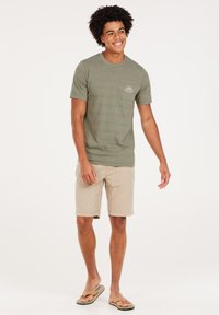 Olijfgroene katoenen T-shirt met korte mouwen en borstzak, gecombineerd met beige shorts en bruine slippers, staande op een effen achtergrond.