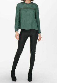 Grüne Bluse mit langen Ärmeln, strukturierten Punktemuster und Spitzenakkzenten, kombiniert mit schwarzen, kunstledernen Hosen und schwarzen, hochhackigen Stiefeletten.