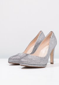 Pár stříbrných třpytivých lodiček na vysokém podpatku s špičatou špičkou vystavených na bílé ploše na světlém pozadí.