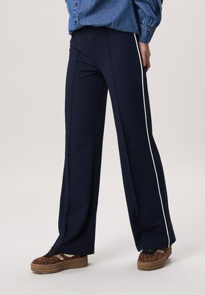 Persoon draagt marineblauwe wijde broek met witte zijstrepen en sneakers met luipaardprint en platformzolen, staand tegen een effen achtergrond.